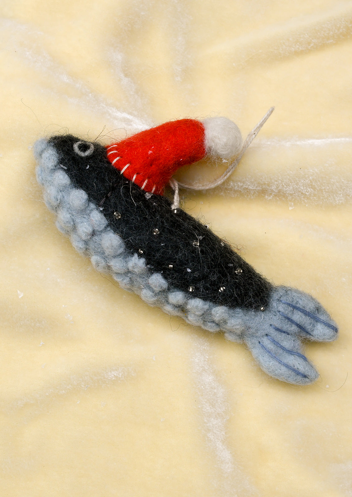 A handcrafted felted wool fish ornament wearing a red Santa hat with a white pom-pom, resting on a textured blue velvet fabric background.