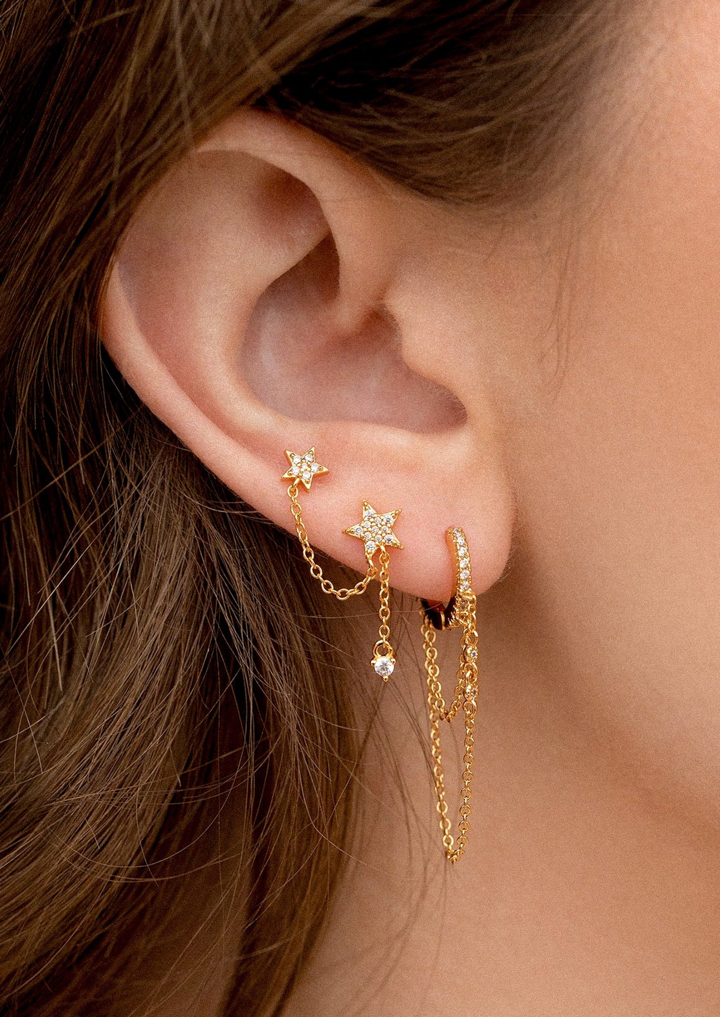 Close-up of an ear wearing gold earrings with star and chain designs.