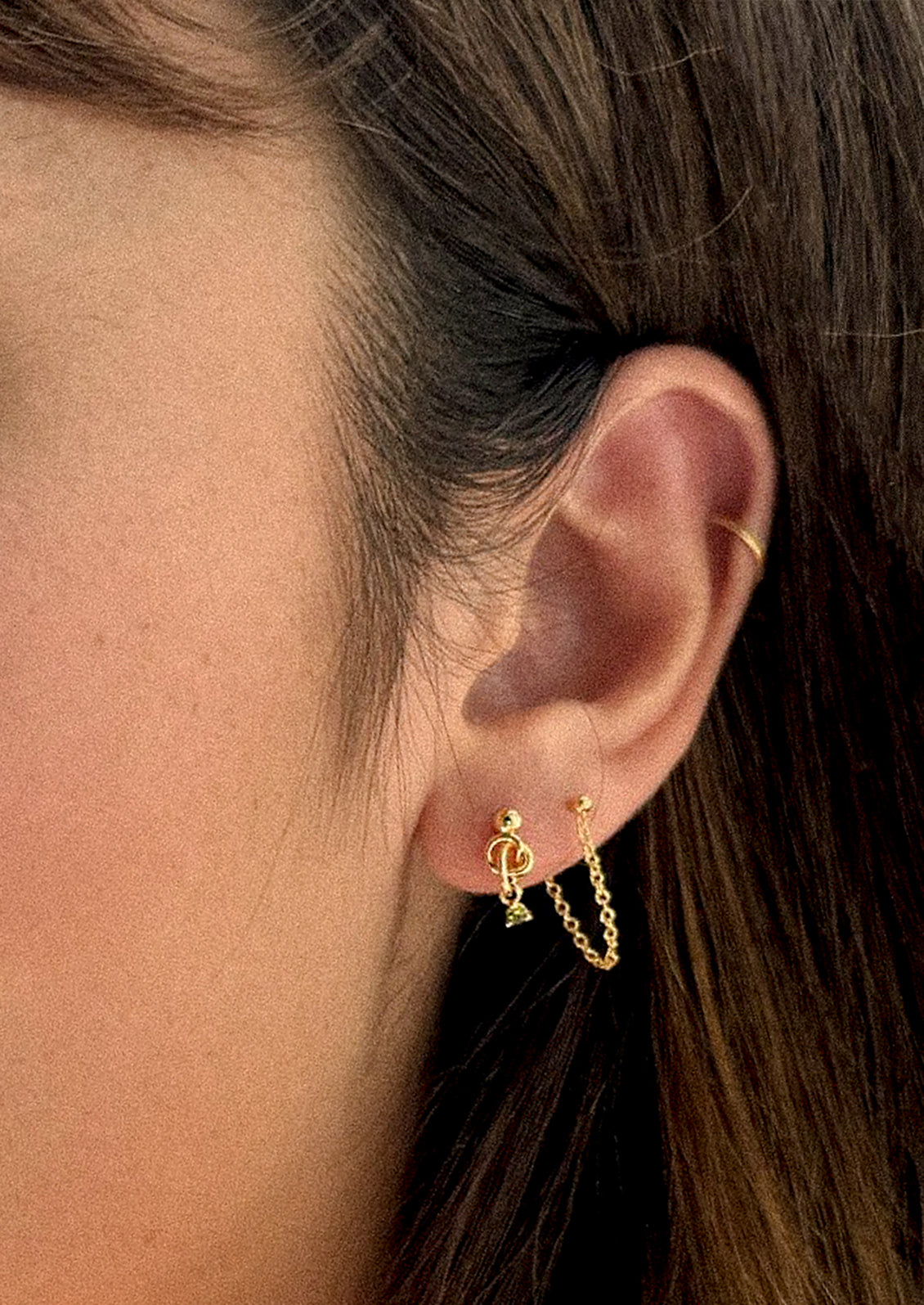 A person wearing a pair of ball stud earrings with knot and green crystal.