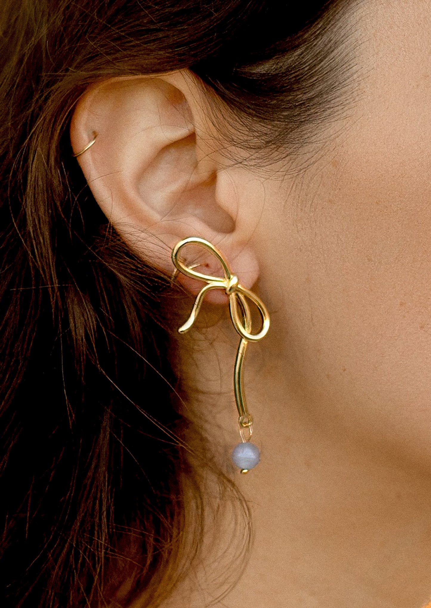 A person wearing a pair of slanted bow earrings in gold with dangling aquamarine gemstone.