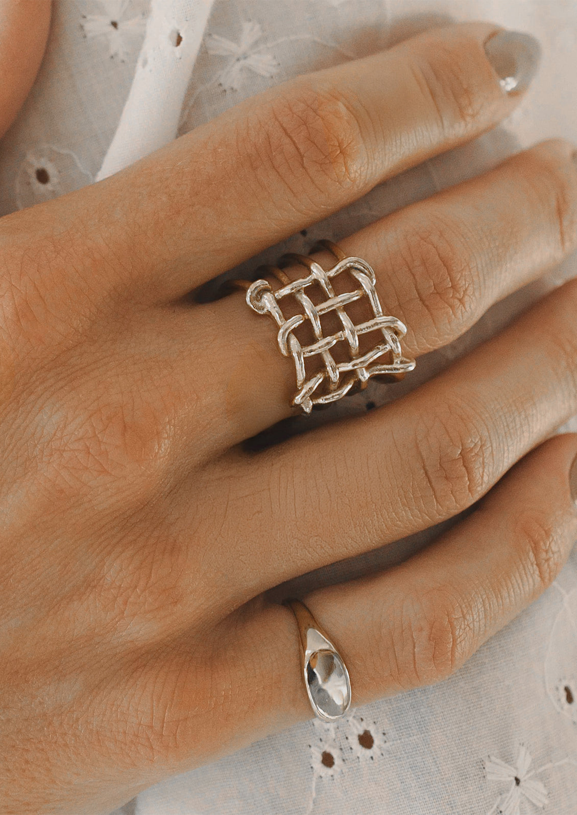 A close-up of a hand wearing a silver ring with a geometric interlaced basketweave design on the middle finger, with a smaller silver signet ring on the ring finger.