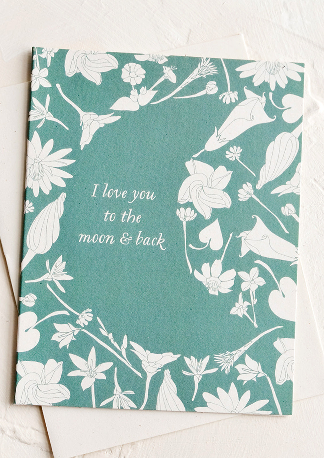 A greeting card with botanicals framed around moon shape, text reads "I love you to the moon & back".
