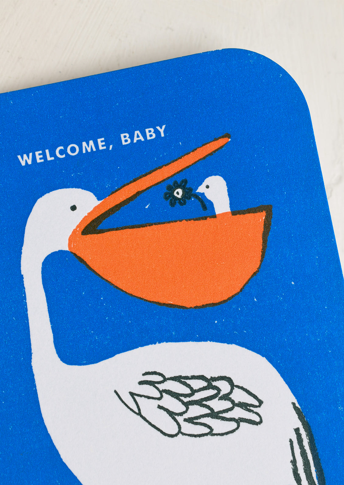 A blue greeting card with illustration of pelican with baby pelican in its beak, text reads "Welcome, baby".