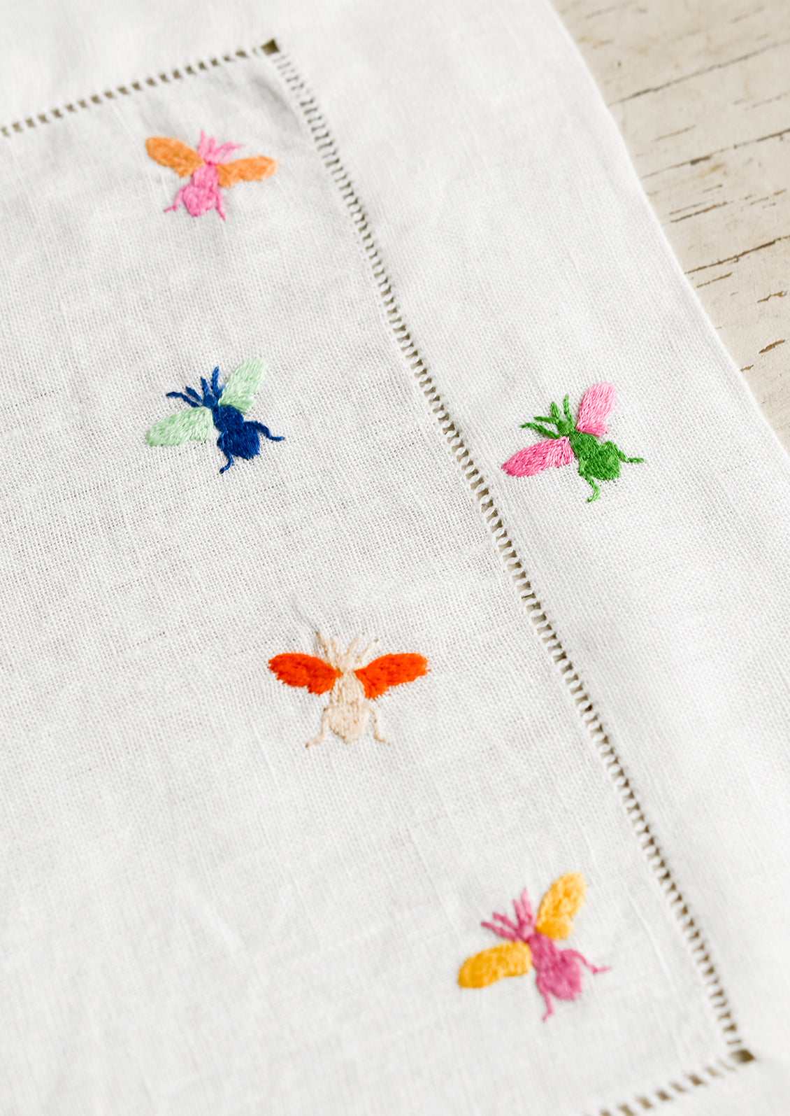 A white linen placemat with multicolor bee embroidery.