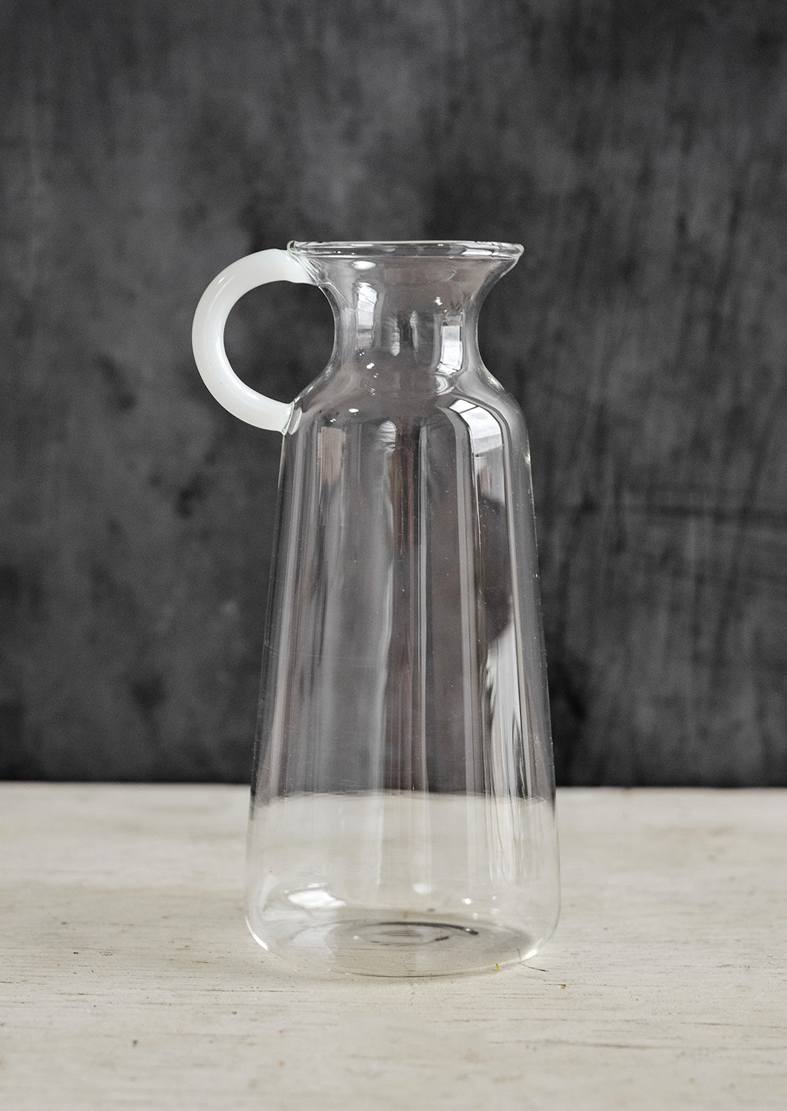 A clear glass vase in milk bottle shape with white handle at one side.