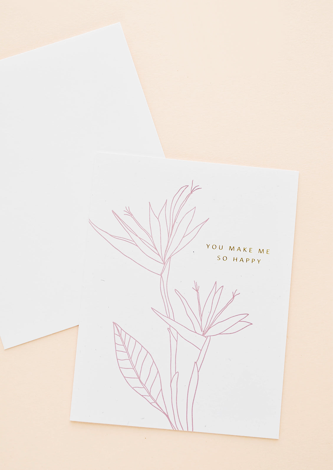 Greeting card with pink flower outline drawing and gold text reading "You make me so happy", with white envelope