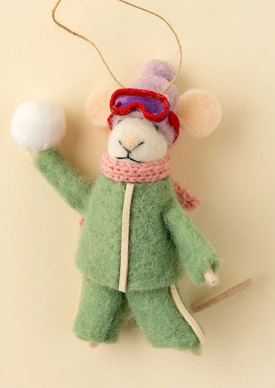 A felted wool mouse ornament dressed in a green ski suit with a pink scarf, red goggles, and a purple hat, holding a small stick against a warm cream background.