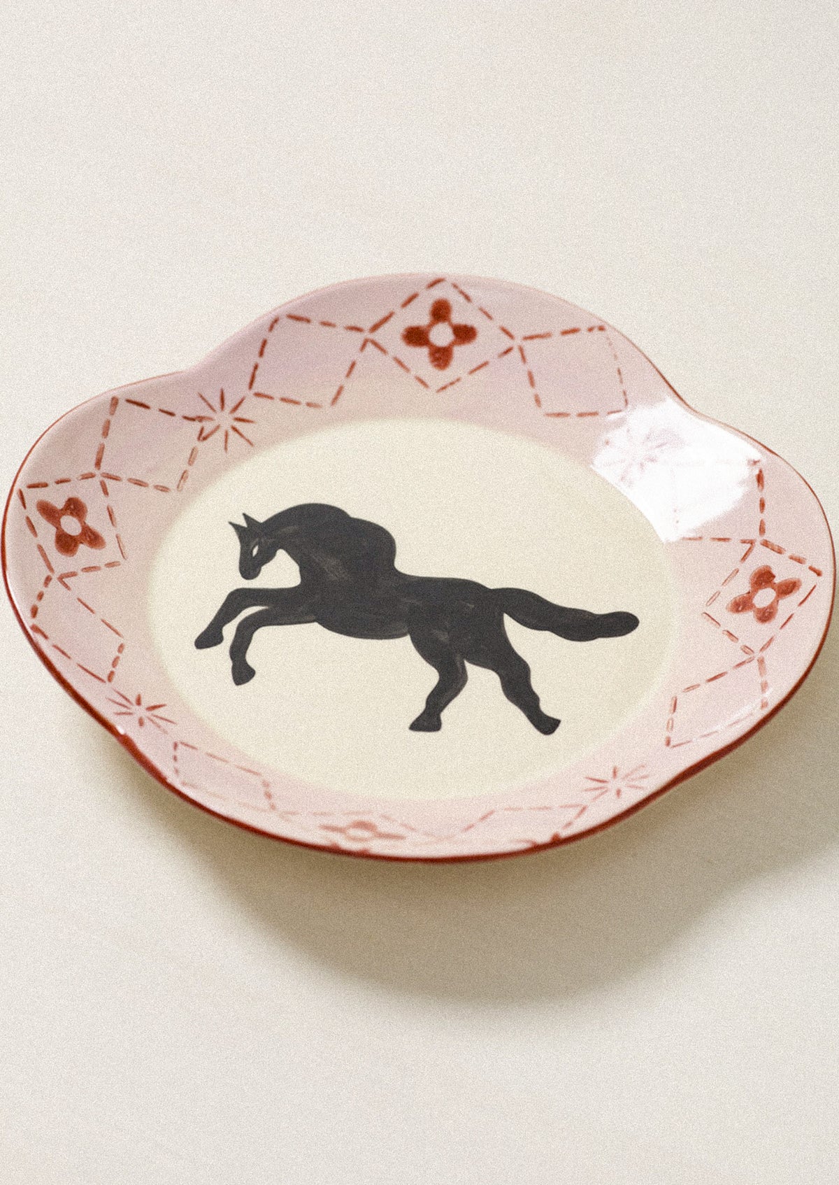 A scalloped ceramic plate with a pink and red patterned rim and a black silhouette of a rearing horse in the center, set against a light beige background.