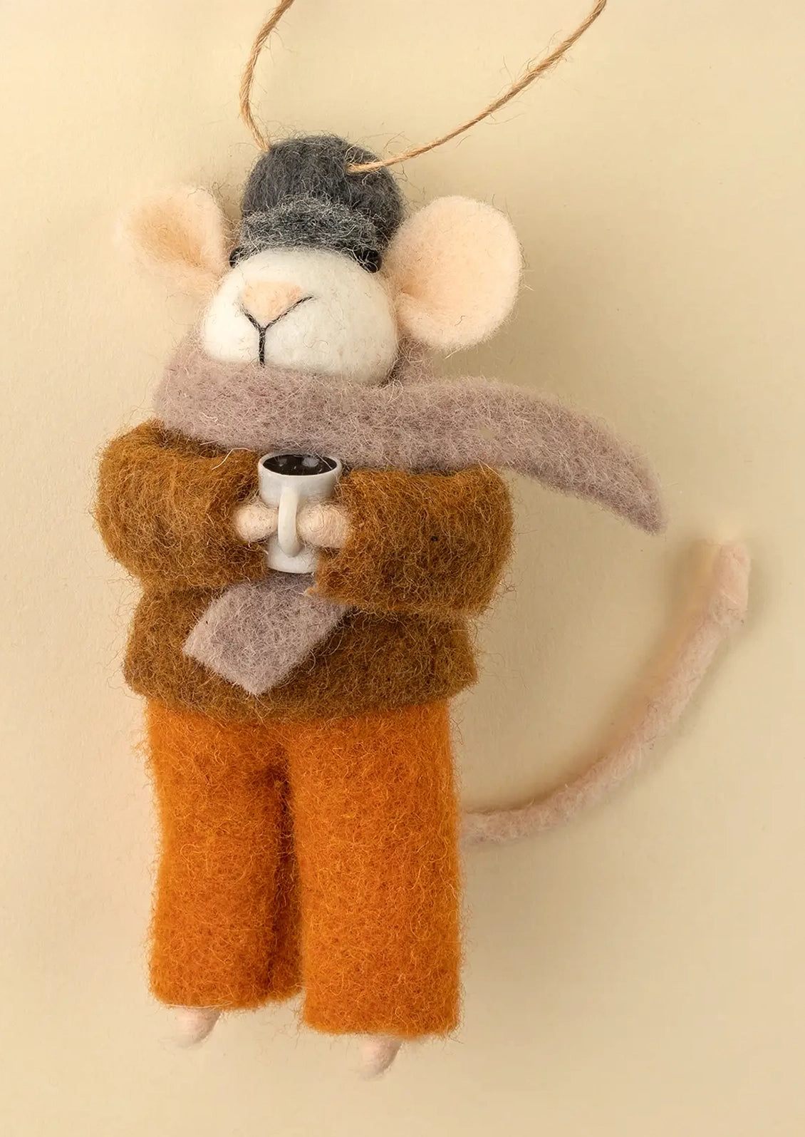 A felted wool mouse ornament wearing a gray beanie, taupe scarf, brown sweater, and orange pants, holding a small white cup, hanging from a twine loop against a warm cream background.