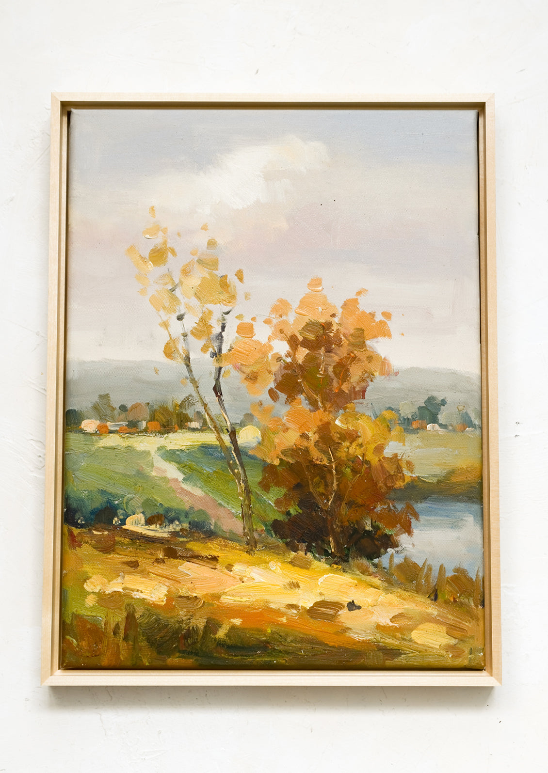 A framed oil landscape painting of two trees next to a pond with houses in the distance.