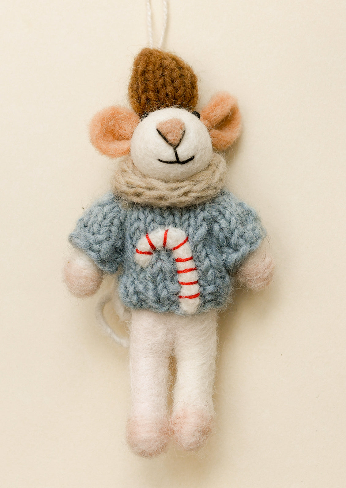 A handmade felted mouse Christmas ornament wearing a blue knit sweater with a red and white candy cane design, a beige scarf, and a brown hat, hanging against a plain cream background.