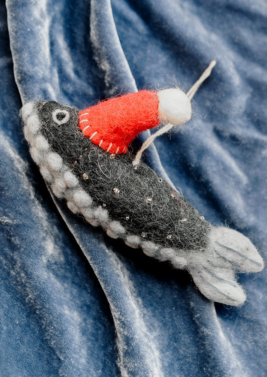 A handcrafted felted wool fish ornament wearing a red knit hat with a white pom-pom, resting on a textured blue velvet fabric background.
