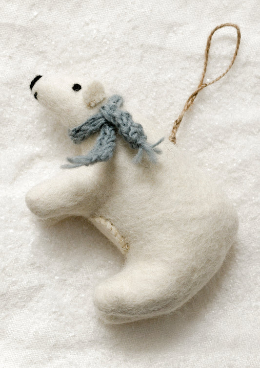 A white felt polar bear Christmas ornament with a knitted blue-gray scarf and a brown jute hanging loop, resting on a textured white background.