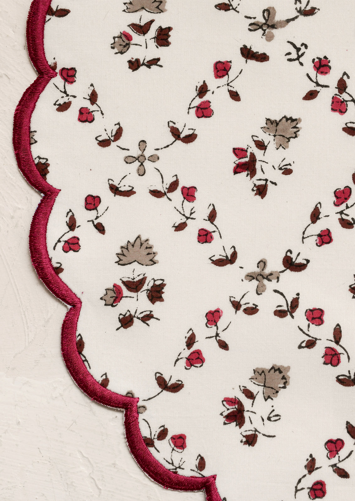 A round scalloped placemat in white with tan and burgundy floral print and embroidered burgundy trim.