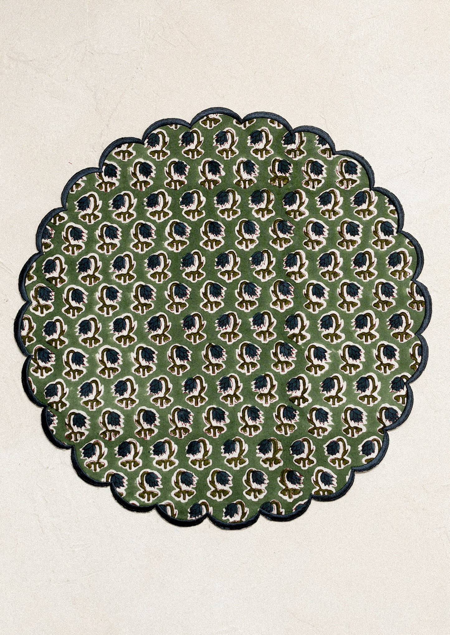 A scalloped round placemat in green with blue floral print and blue embroidered trim.