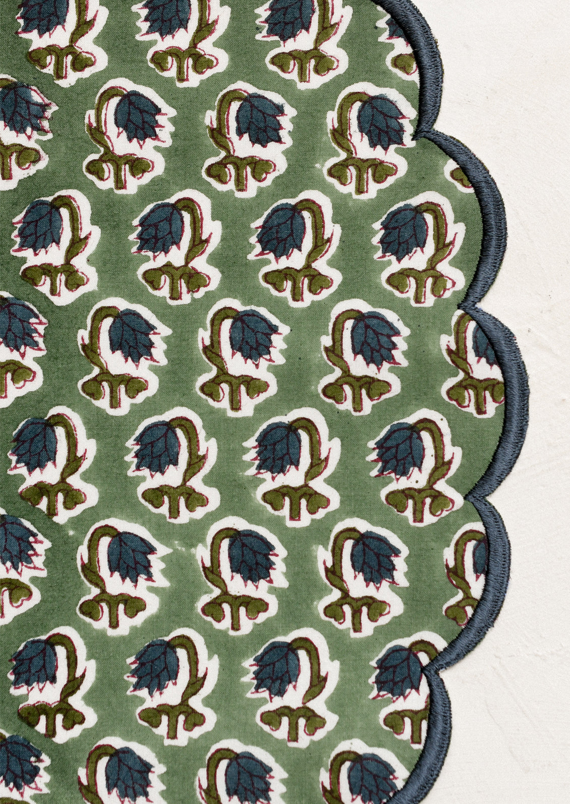 A scalloped round placemat in green with blue floral print and blue embroidered trim.