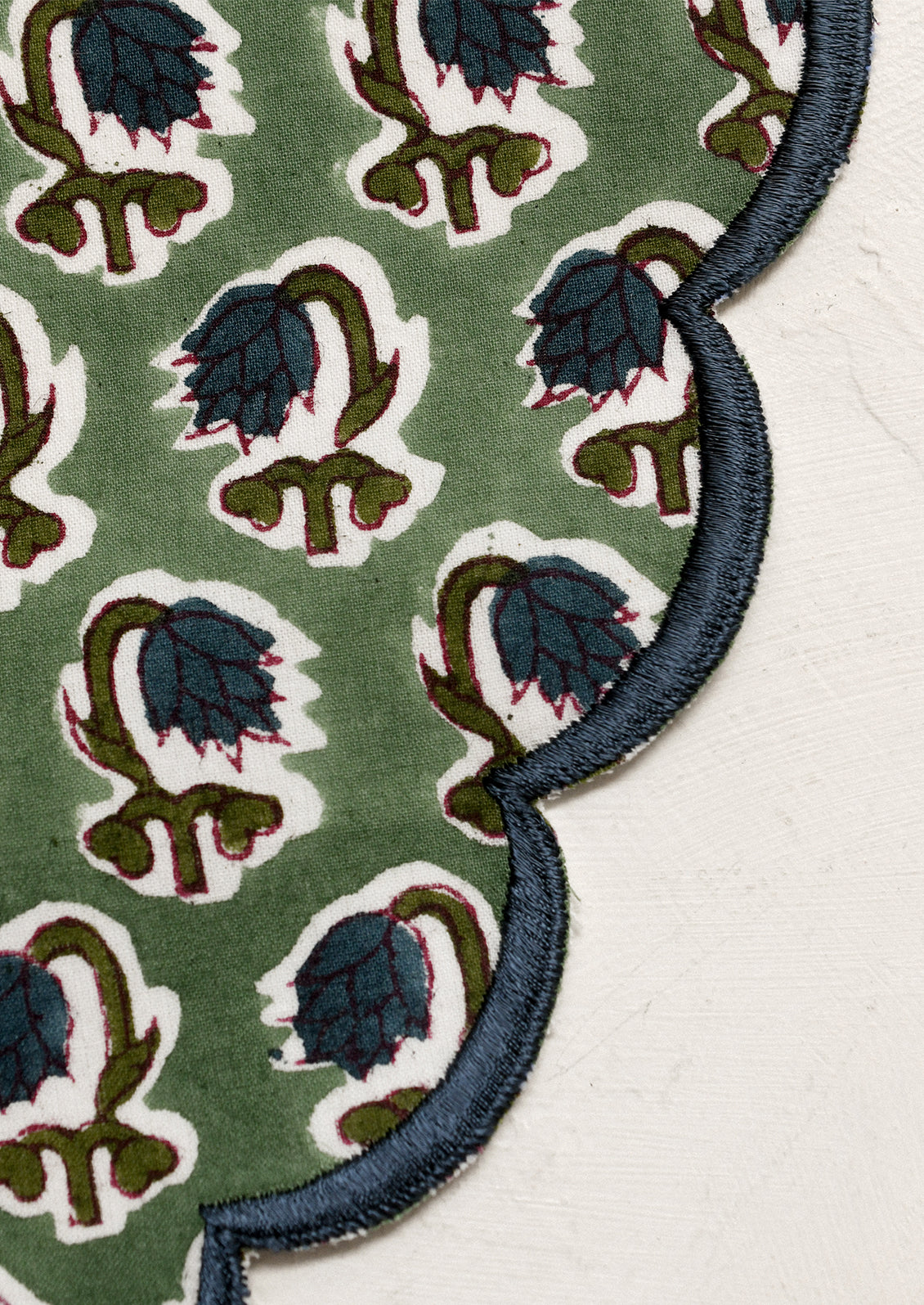 A scalloped round placemat in green with blue floral print and blue embroidered trim.