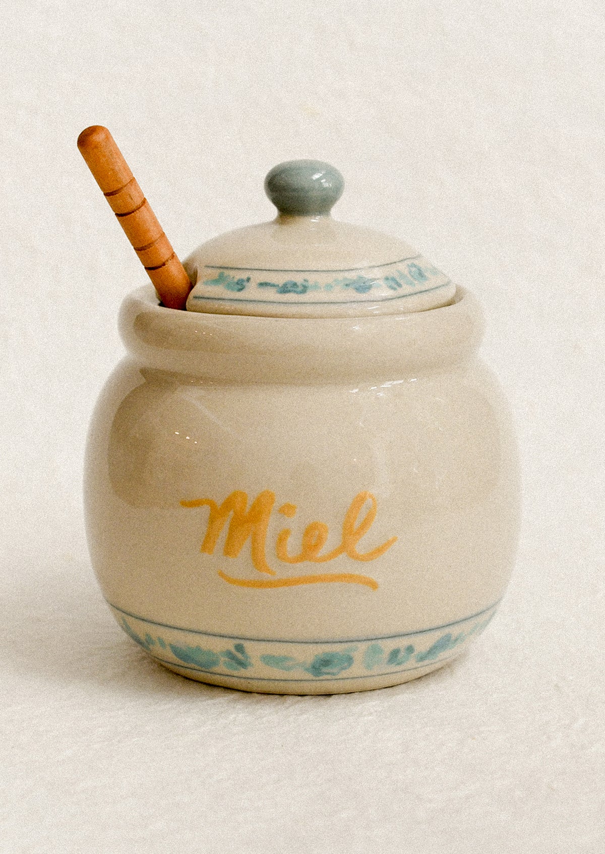 A stoneware honey jar with "Miel" written in yellow script, with blue floral stripe trim.