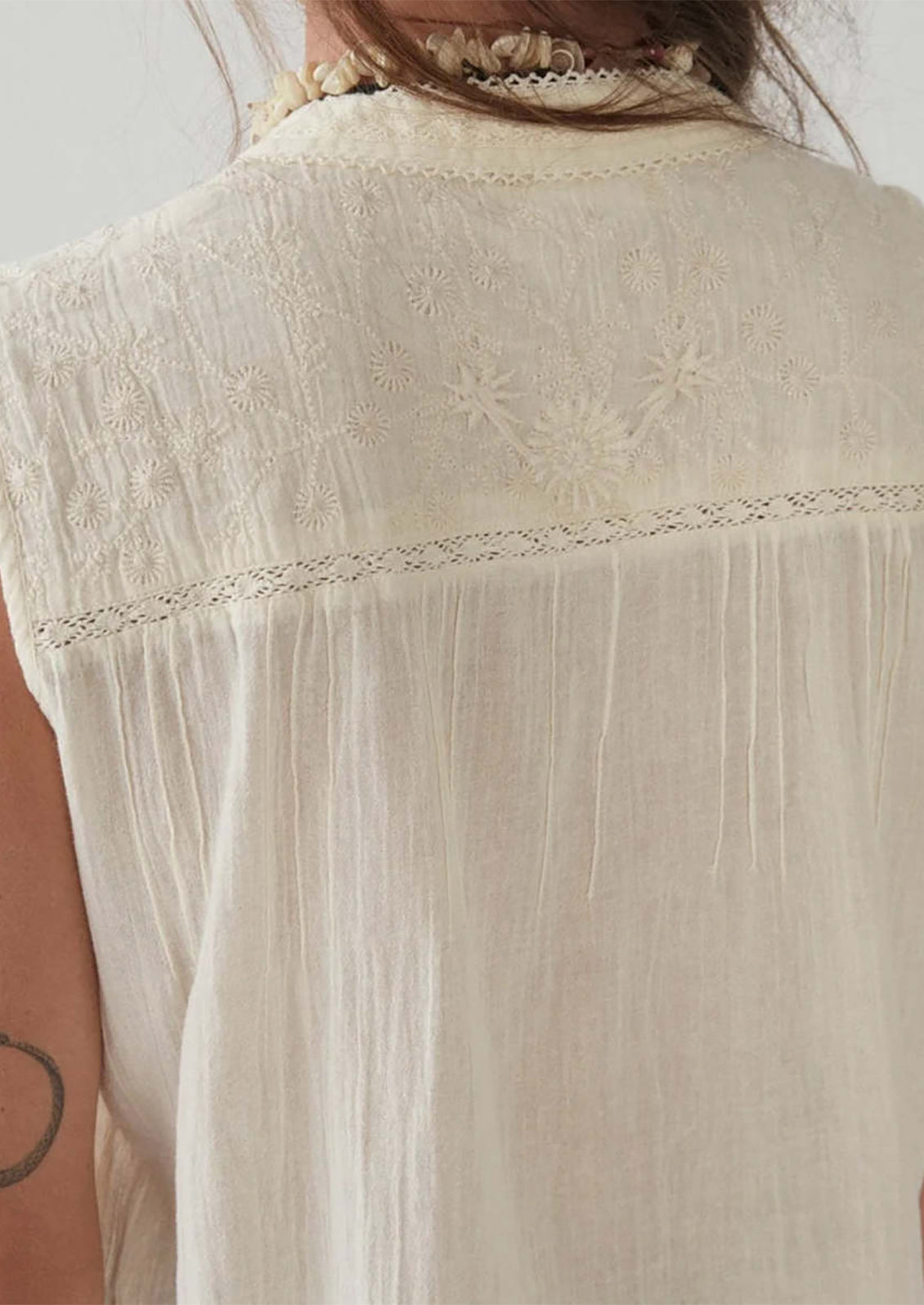 A woman wearing a sleeveless cotton button front blouse with tonal embroidered floral pattern at yoke.