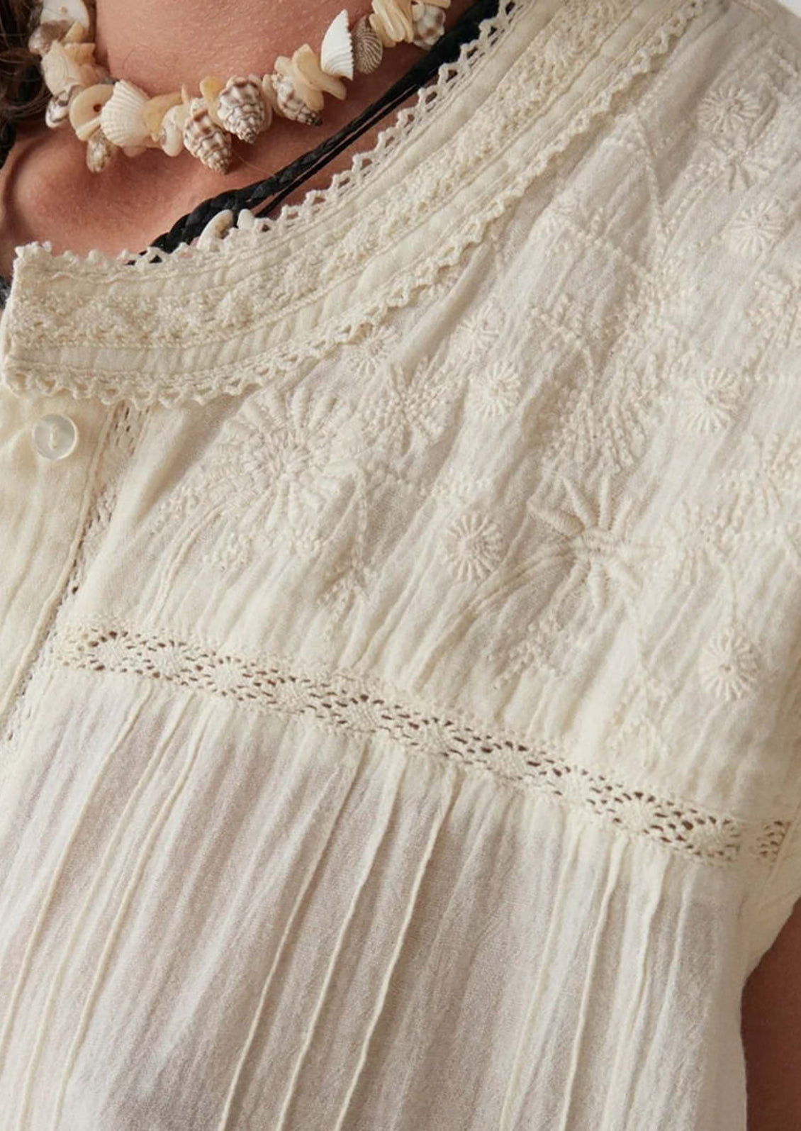 A woman wearing a sleeveless cotton button front blouse with tonal embroidered floral pattern at yoke.