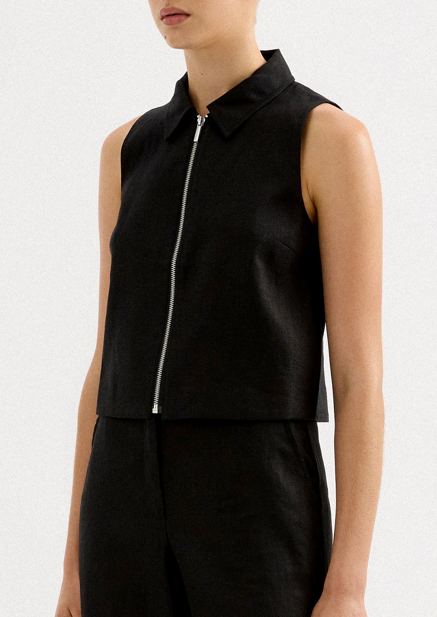 A woman wearing a sleeveless collared shirt with full length silver zipper at front placket.