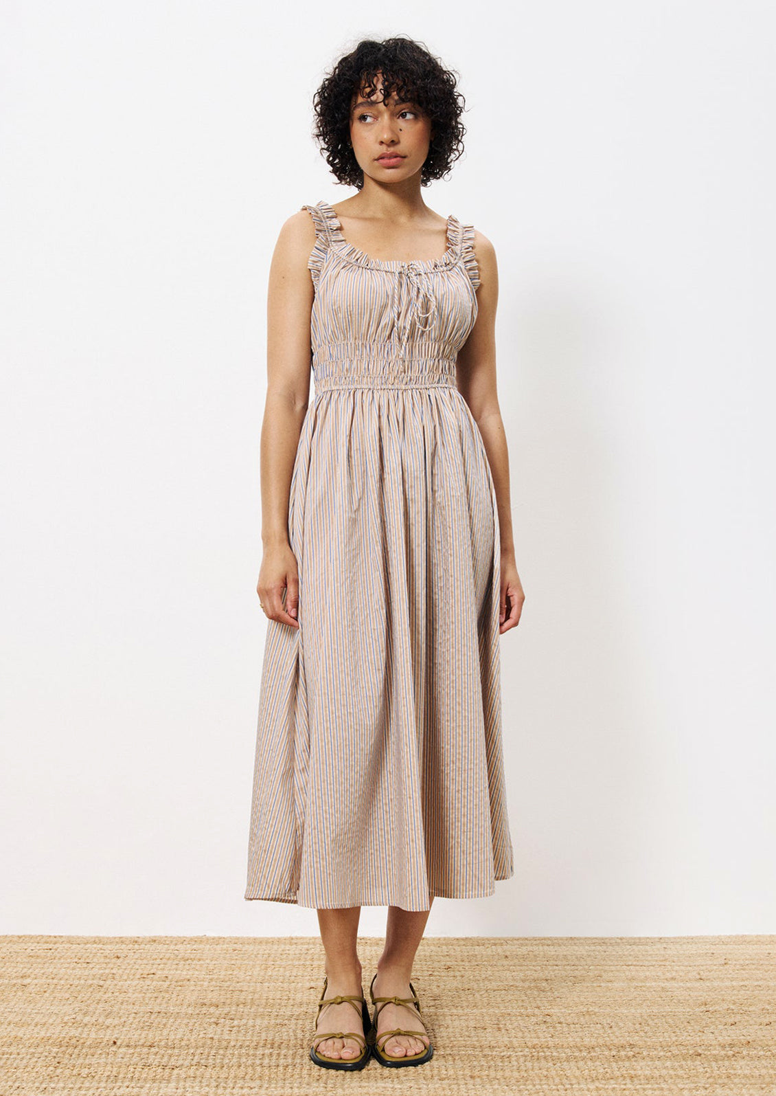 A woman wearing a smocked waist dress with ruffle trim, in brown and blue pinstripe.