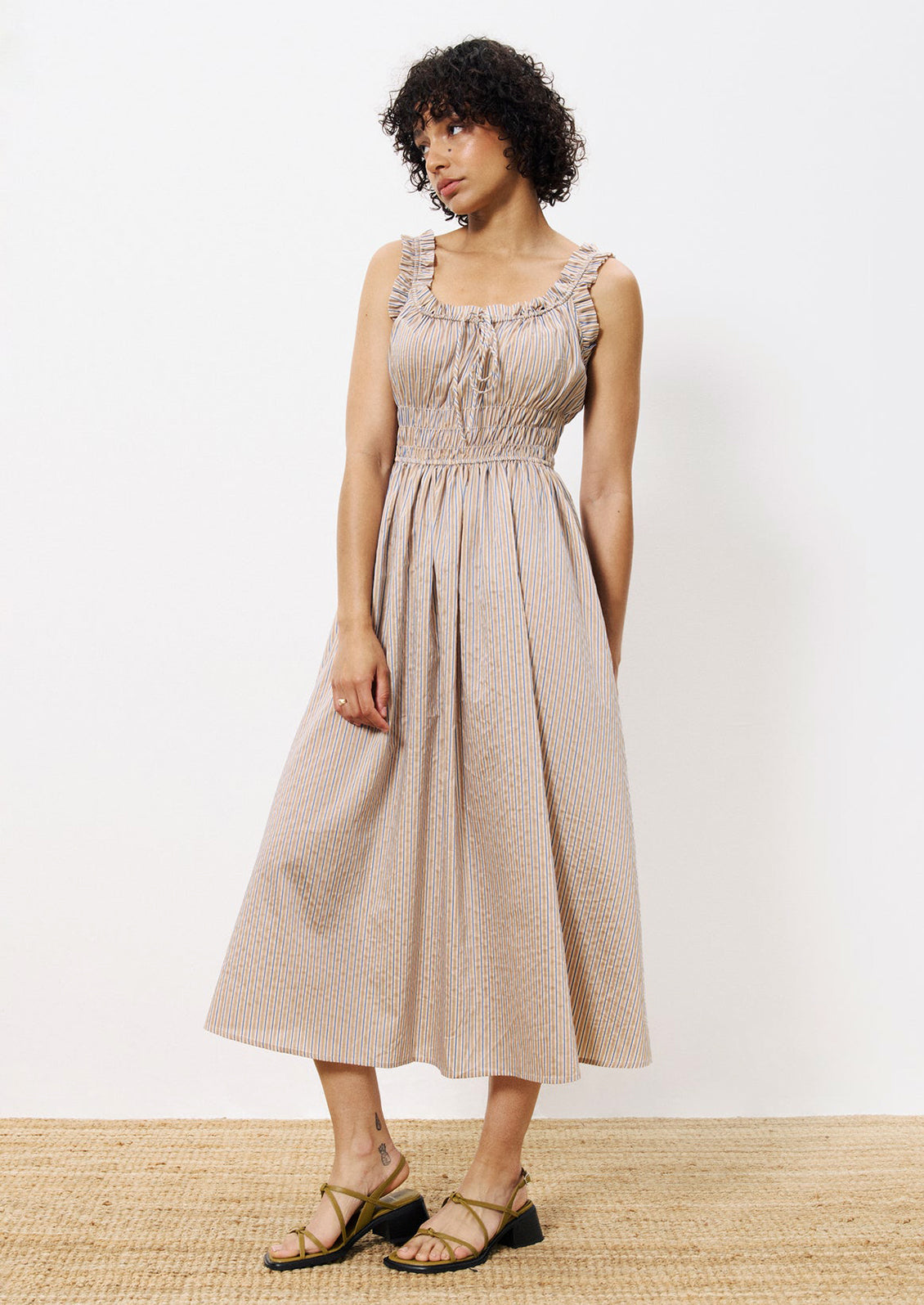 A woman wearing a smocked waist dress with ruffle trim, in brown and blue pinstripe.