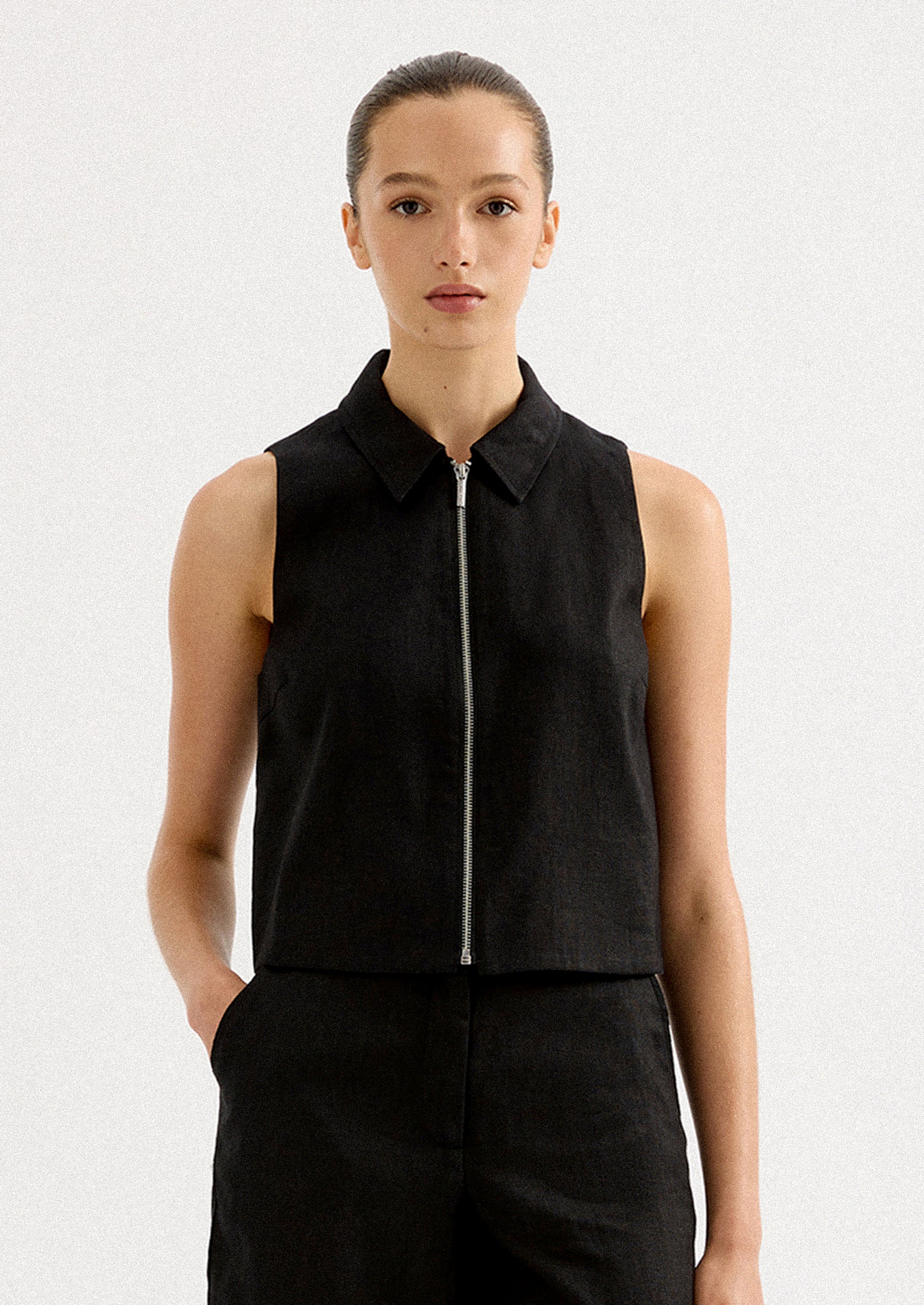 A woman wearing a sleeveless collared shirt with full length silver zipper at front placket.