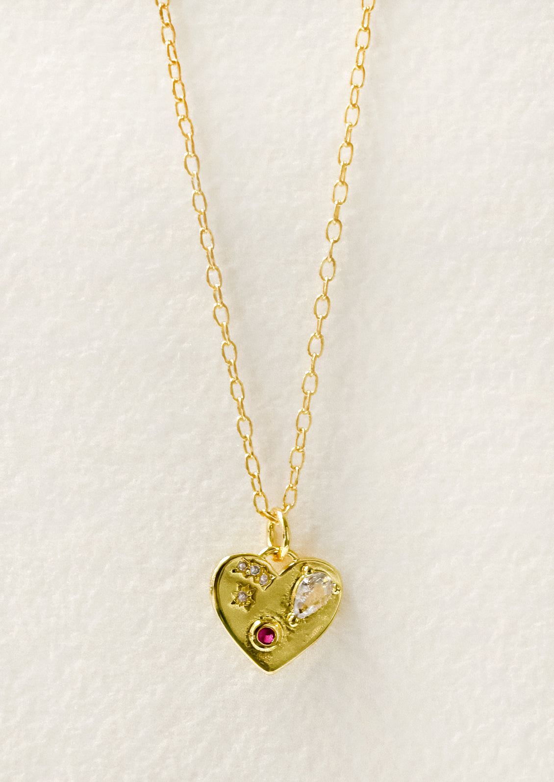 A gold necklace with a gold heart charm adorned with glass crystal and a red gemstone.