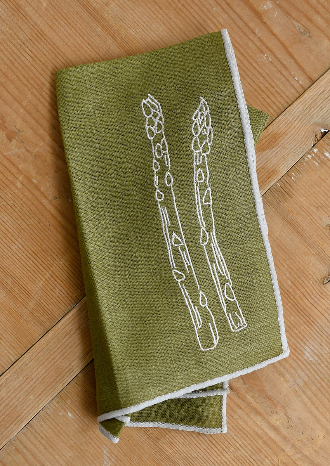 An olive green cloth dinner napkin with grey edging, and grey embroidery of asparagus spears.