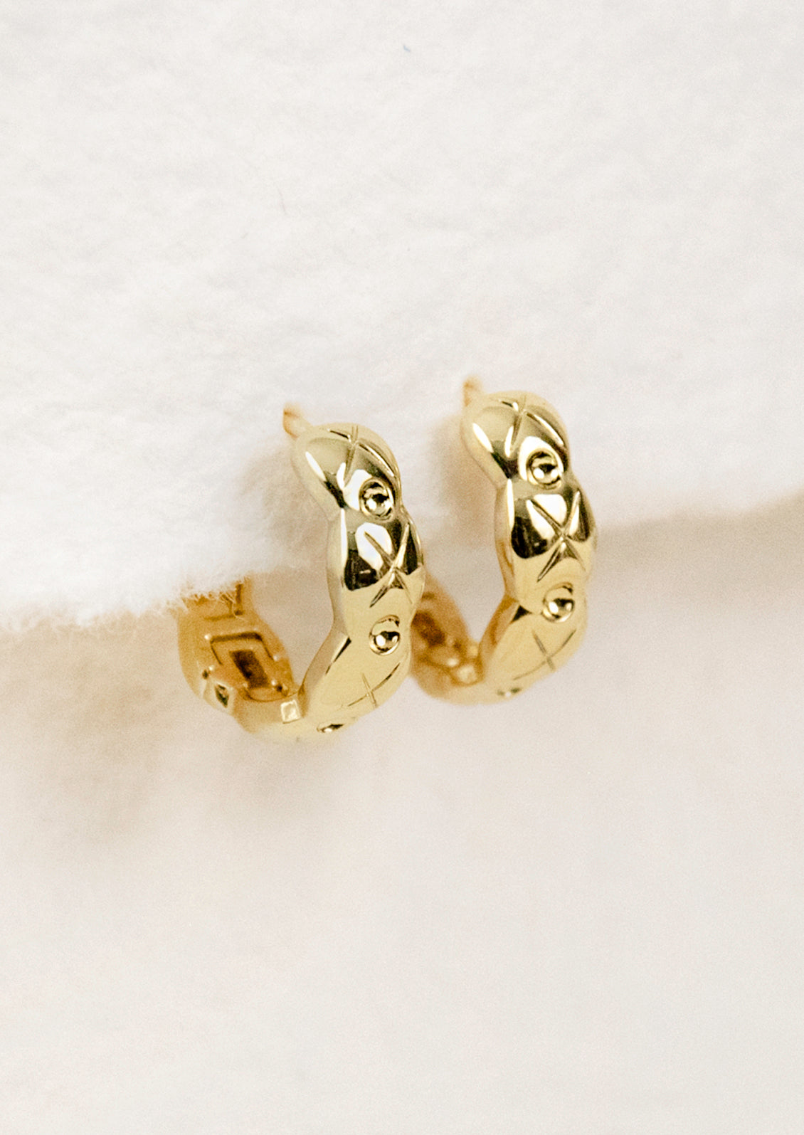 A pair of gold huggie hoop earrings with wavy edges and an etched design.