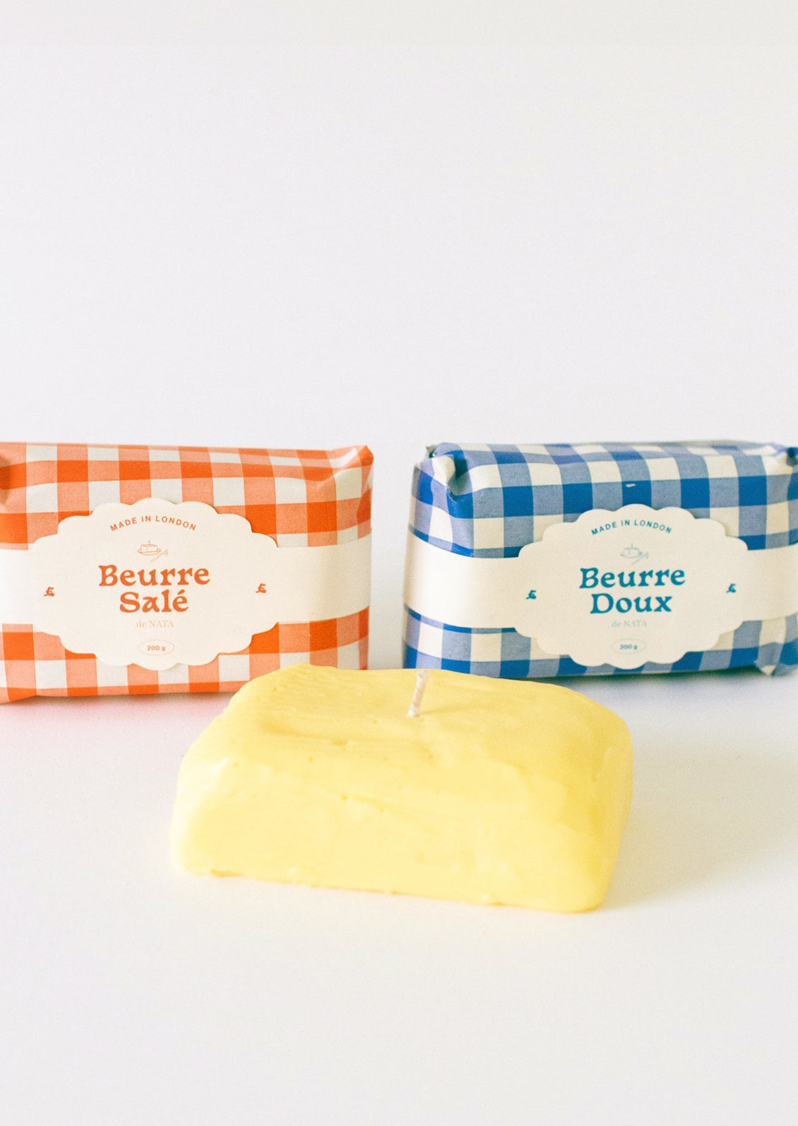 A candle in the shape of a block of butter, gingham packaging in red or blue.
