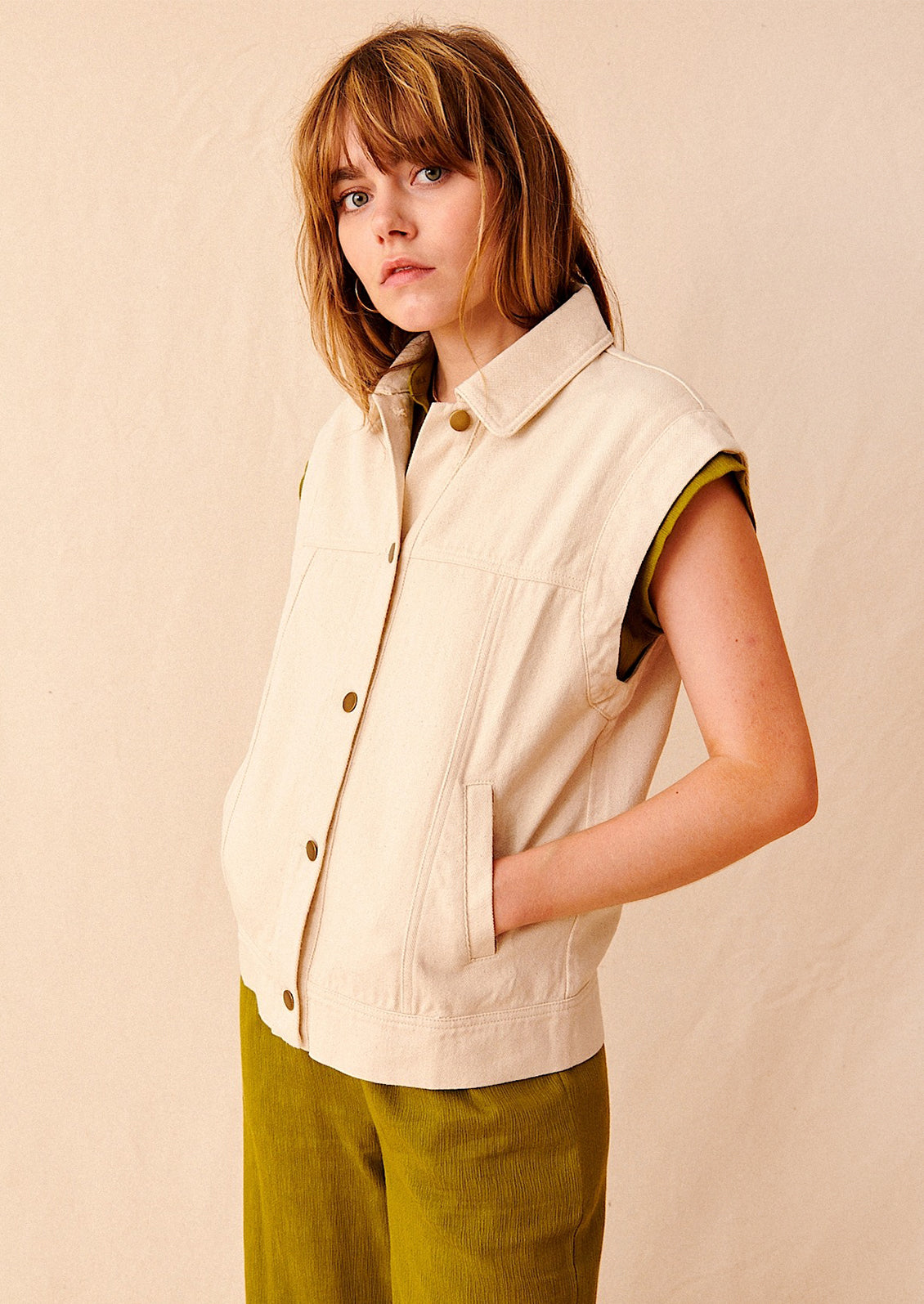 A woman wearing an ecru-colored sleeveless denim jacket. 