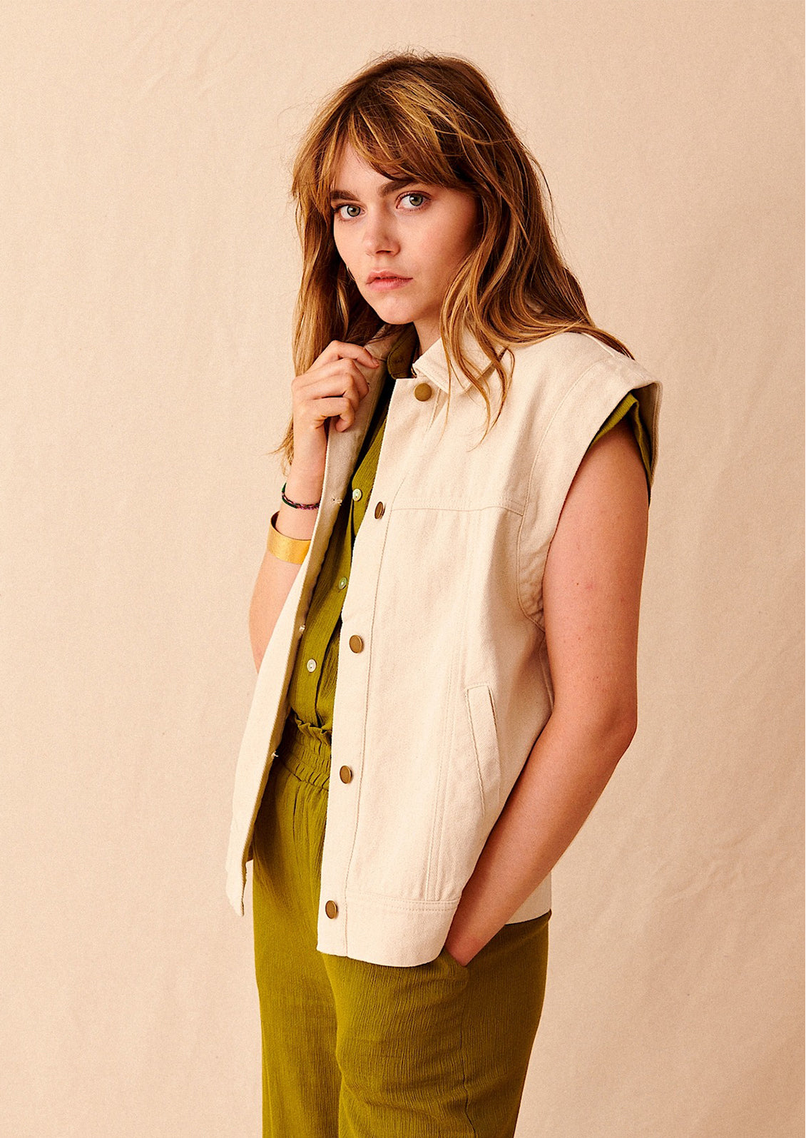 A woman wearing an ecru-colored sleeveless denim jacket over green shirt and pants. 