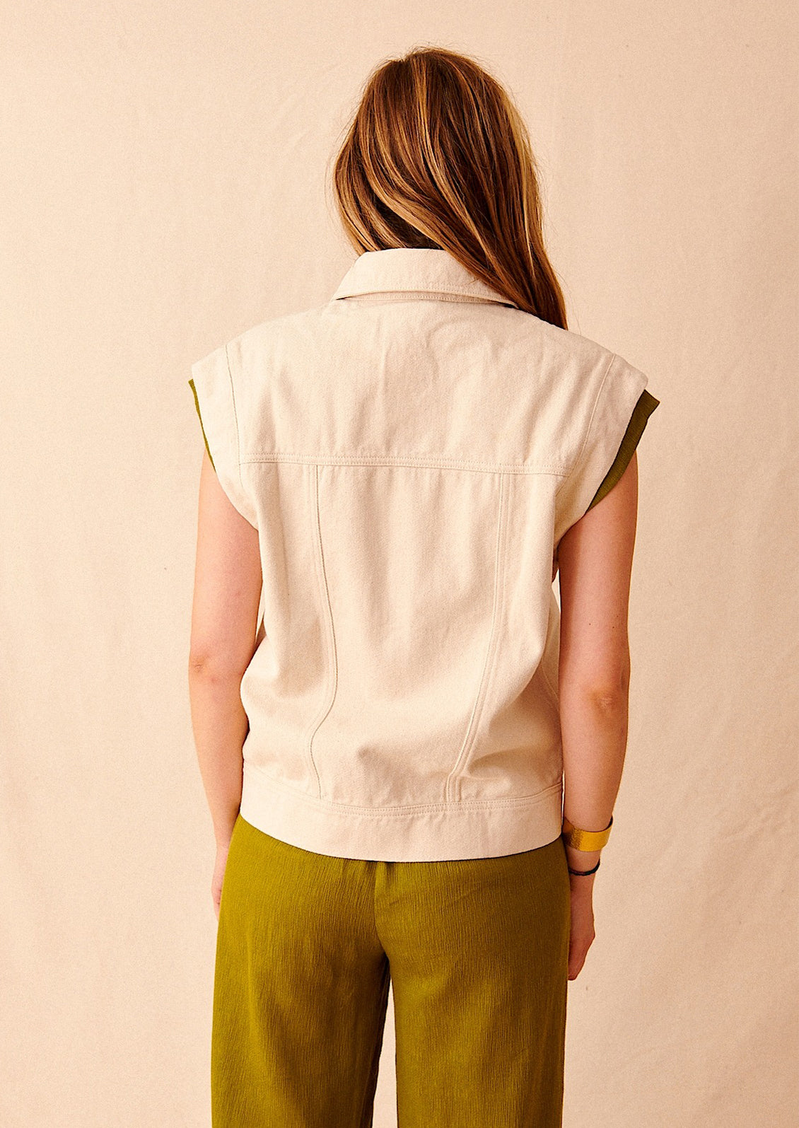 A woman wearing an ecru-colored sleeveless denim jacket and green pants, seen from behind. 