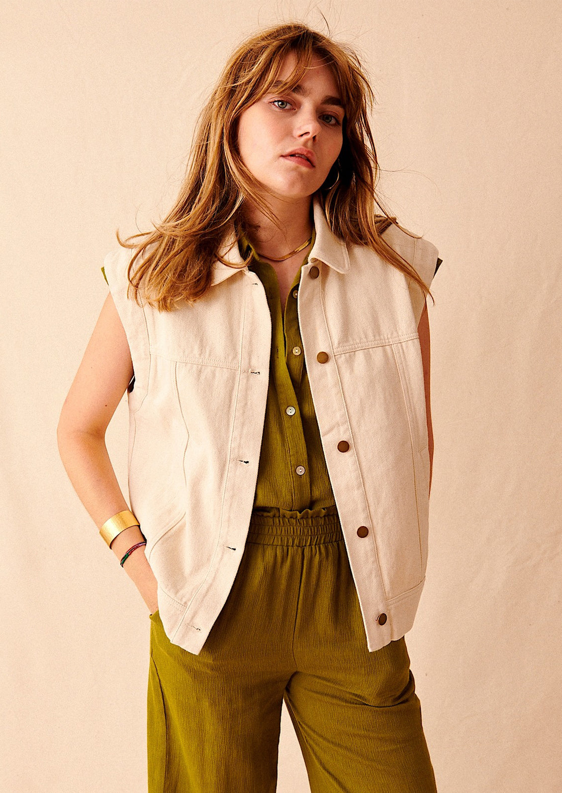A woman wearing an ecru-colored sleeveless denim jacket over green shirt and pants. 