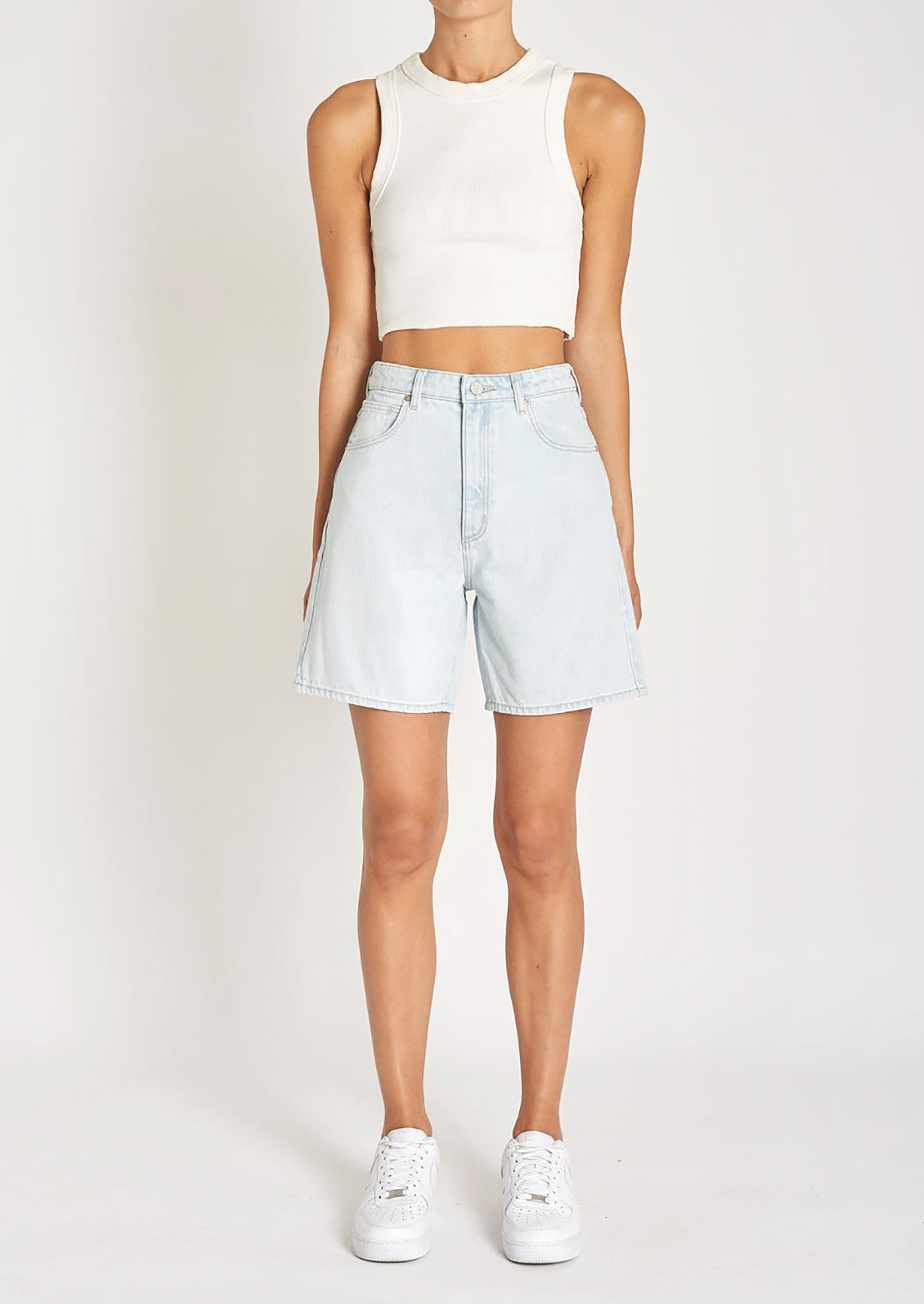 A woman in a white tank top, light-wash denim shorts, and sneakers. 