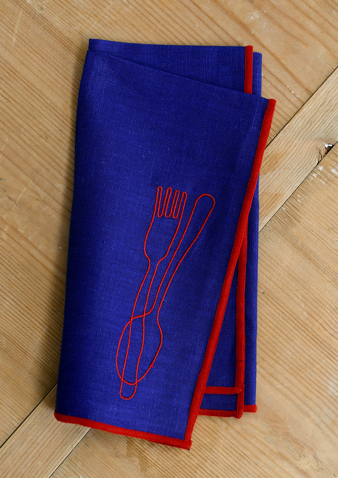 A royal blue cloth dinner napkin with red edging, and red embroidery of a spoon and fork.