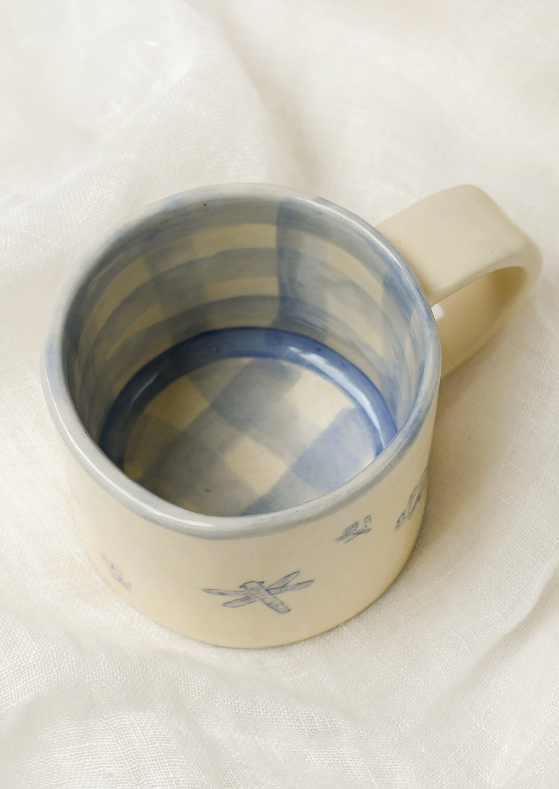 Top view of a handmade ceramic mug with illustrations of flowers and tiny insects on the outside and a gingham pattern on the inside.