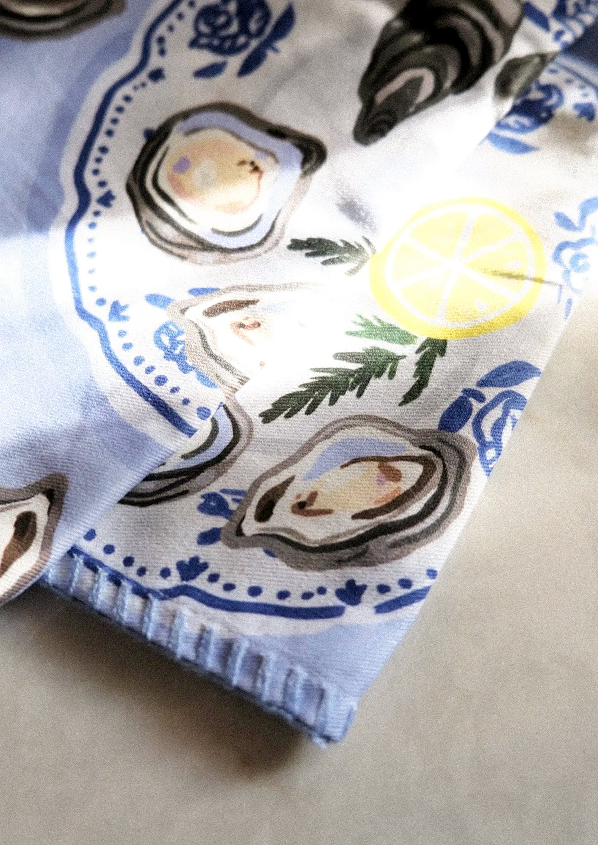 A printed tea towel with illustration of oysters and lemon wedges on periwinkle background.