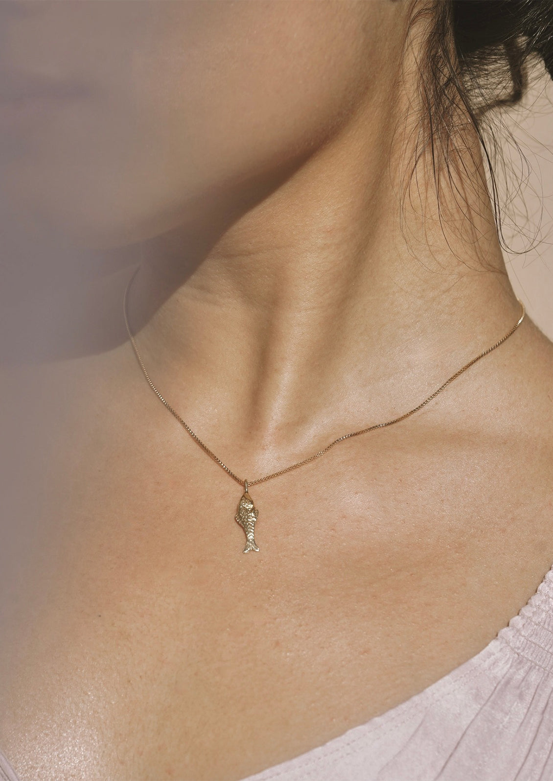 A person wearing a gold necklace with fish charm on thin box chain.