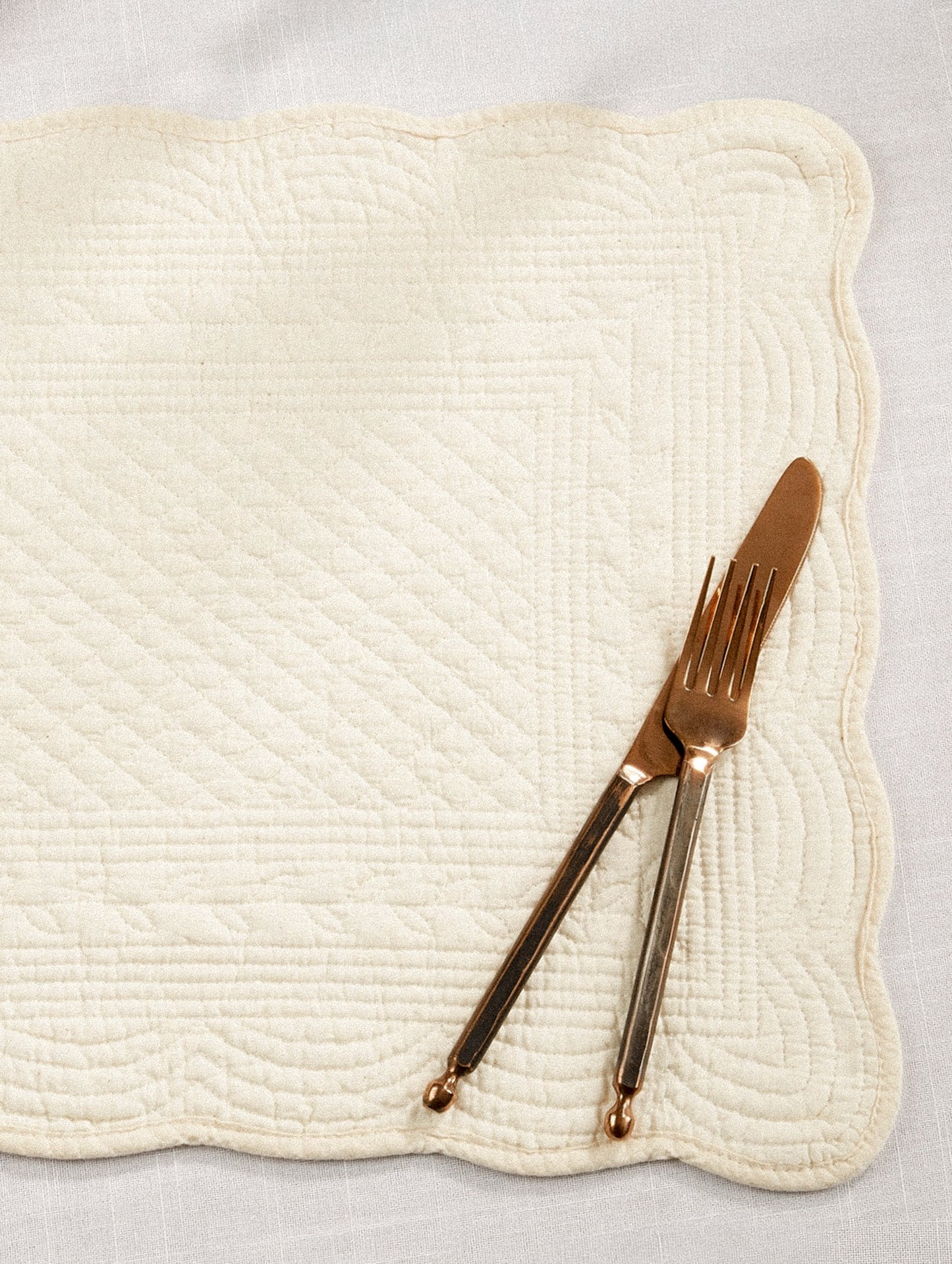 A quilted cotton placemat with scalloped edges, in ivory.