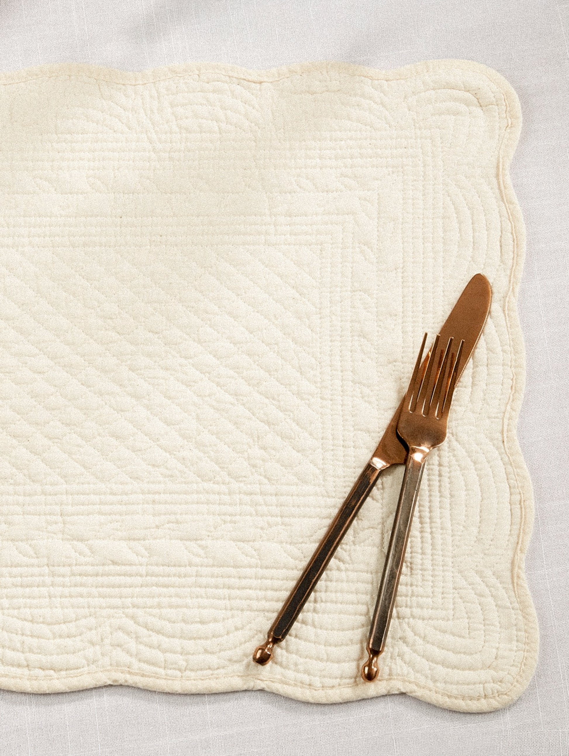 A quilted cotton placemat with scalloped edges, in ivory.