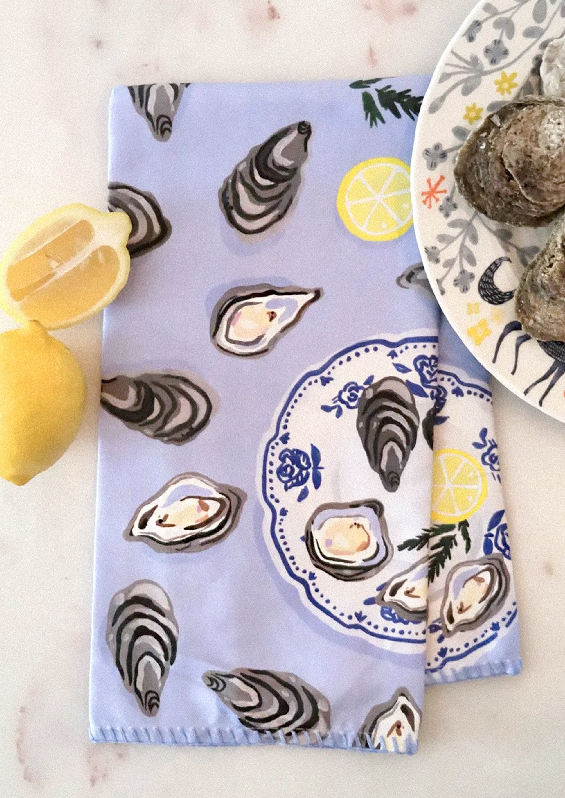 A printed tea towel with illustration of oysters and lemon wedges on periwinkle background.