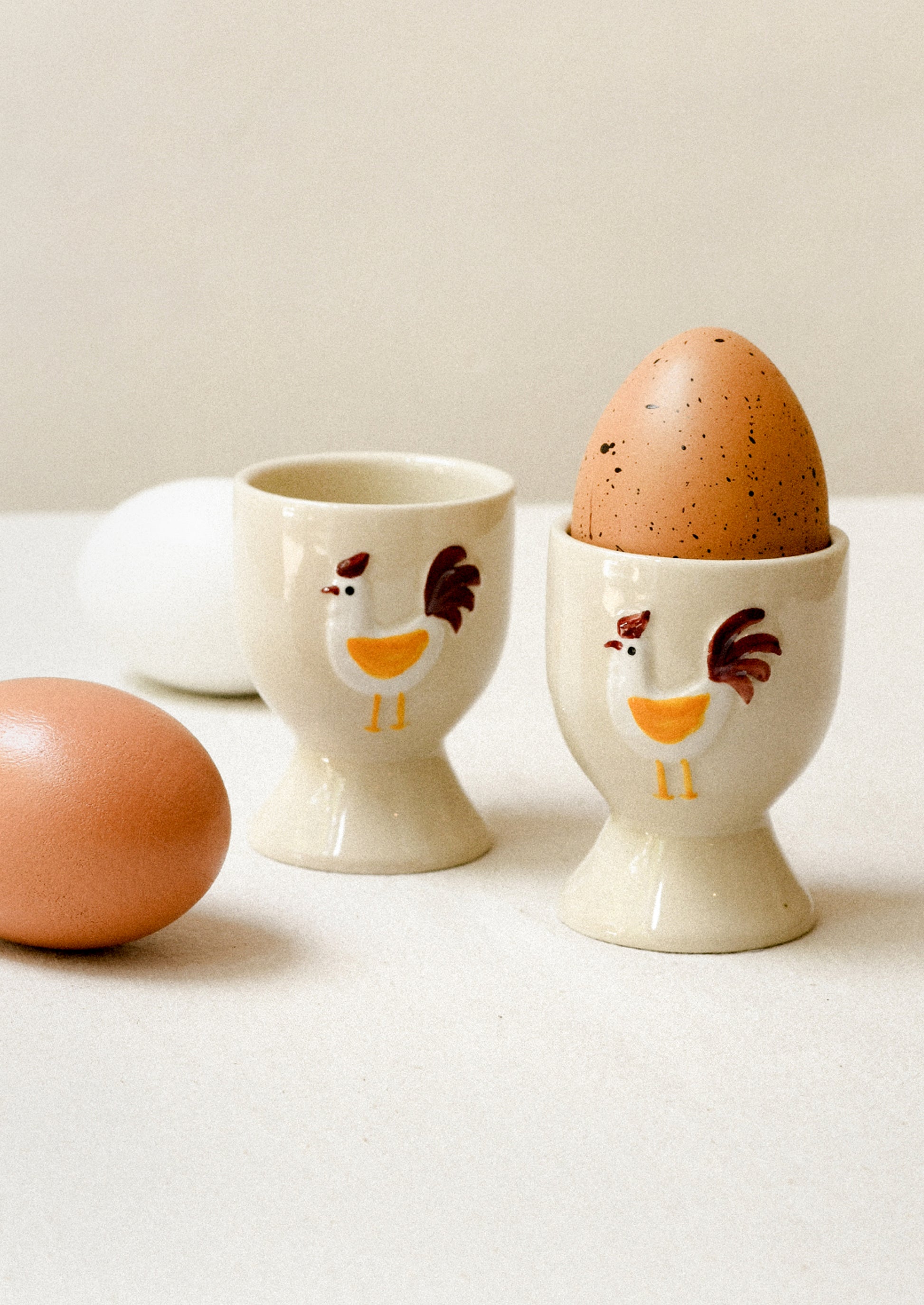 Two ceramic egg cups with chicken designs on a white surface, containing eggs.