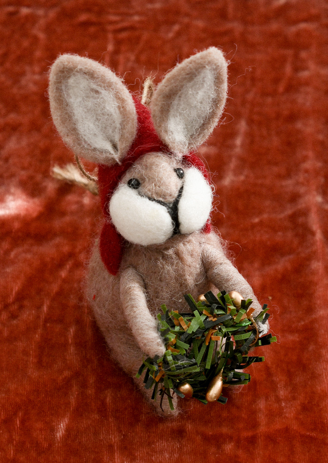 A felted beige bunny ornament wearing red hood and carrying wreath.