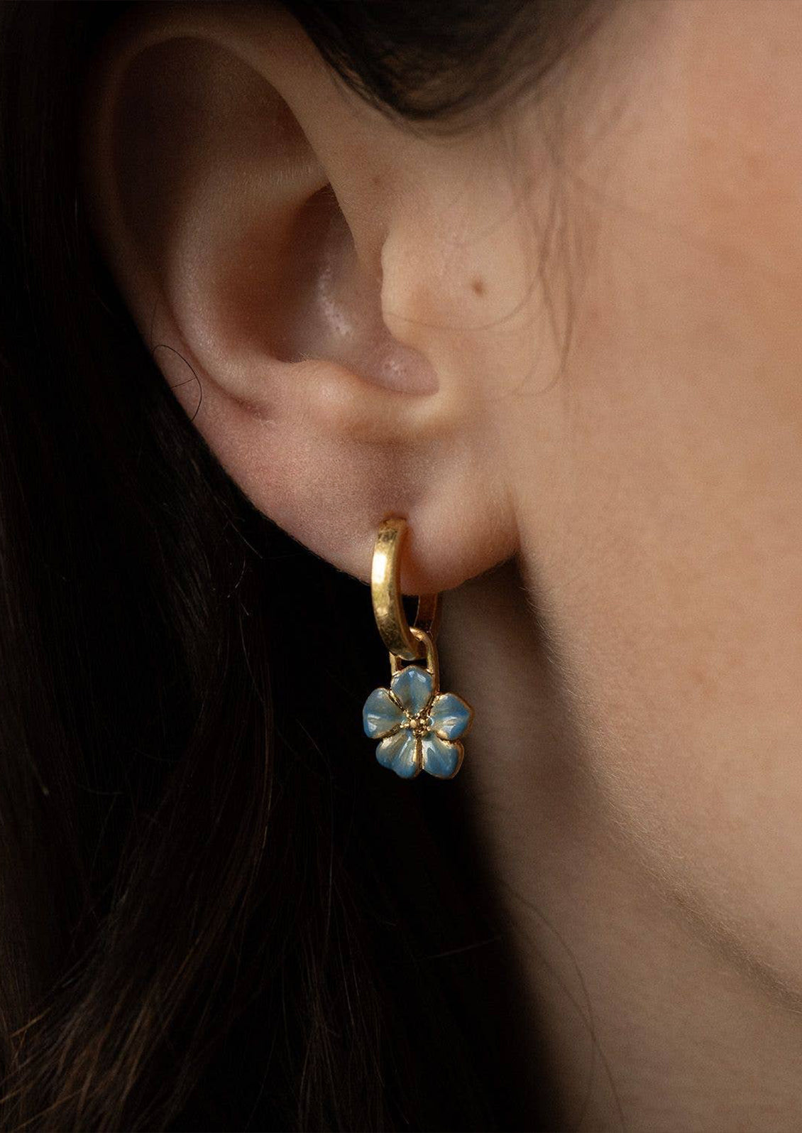 A woman wearing a gold hoop earring with a blue forget me not flower charm.