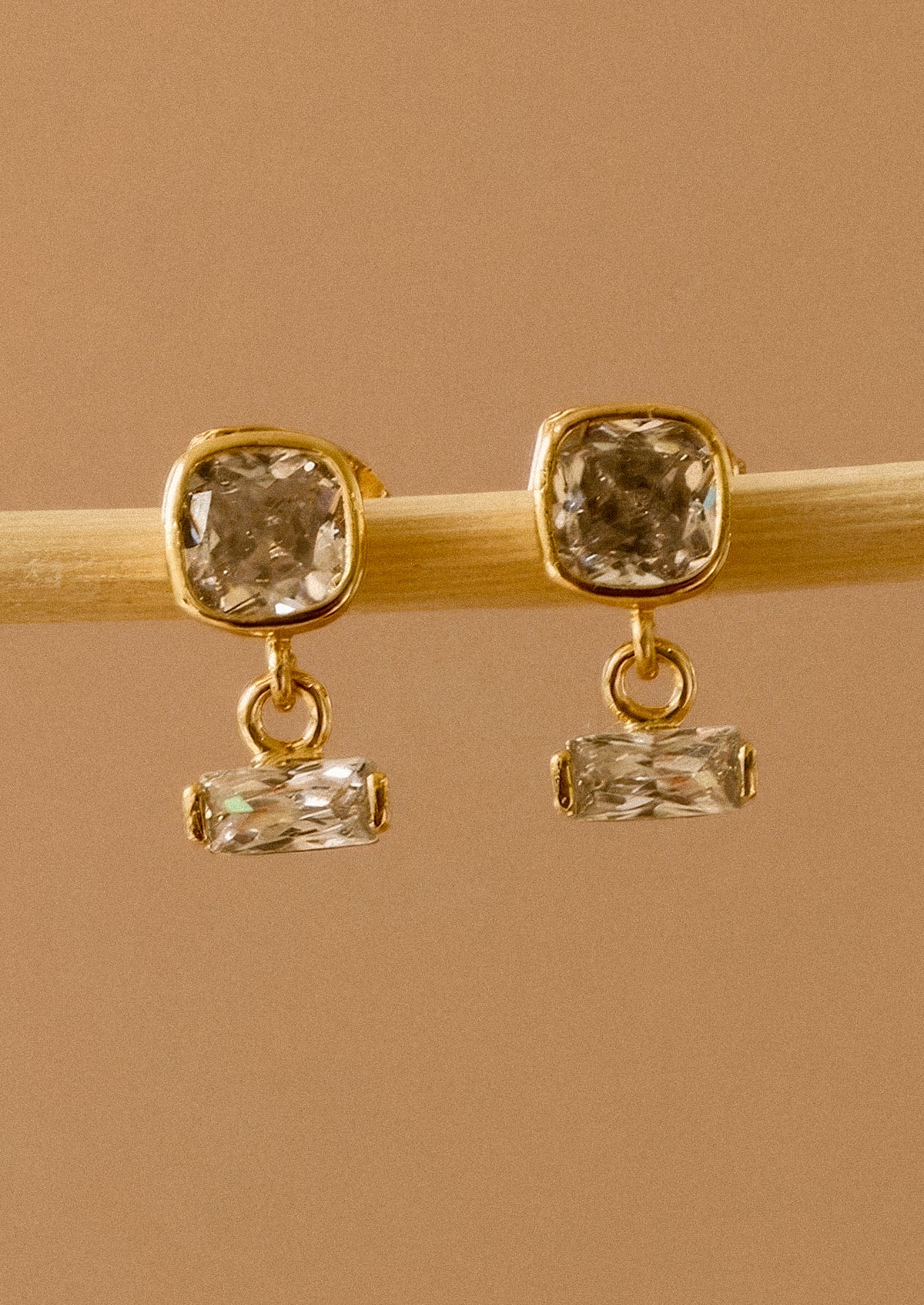 A pair of gold drop earrings with square cushion-cut clear crystals on the top and smaller rectangular clear crystals below, displayed on a light wooden stick against a warm tan background.