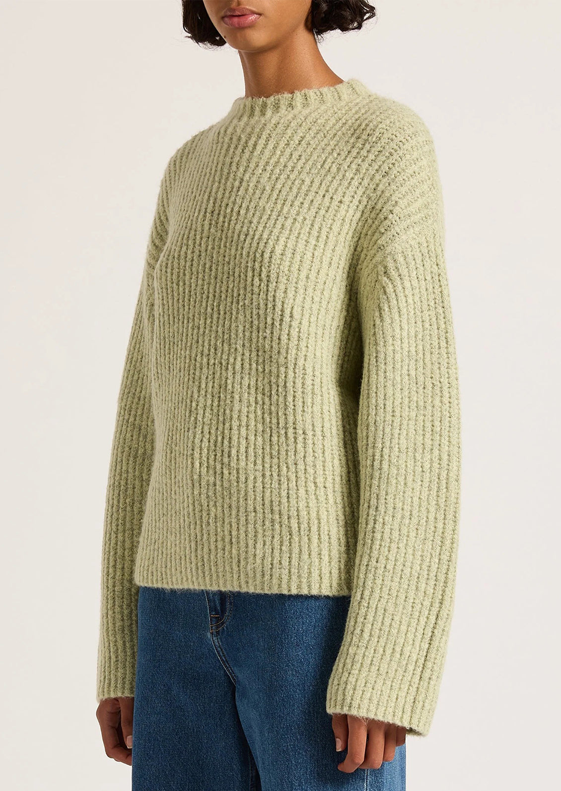 Person wearing a light green ribbed sweater and blue jeans on a white background