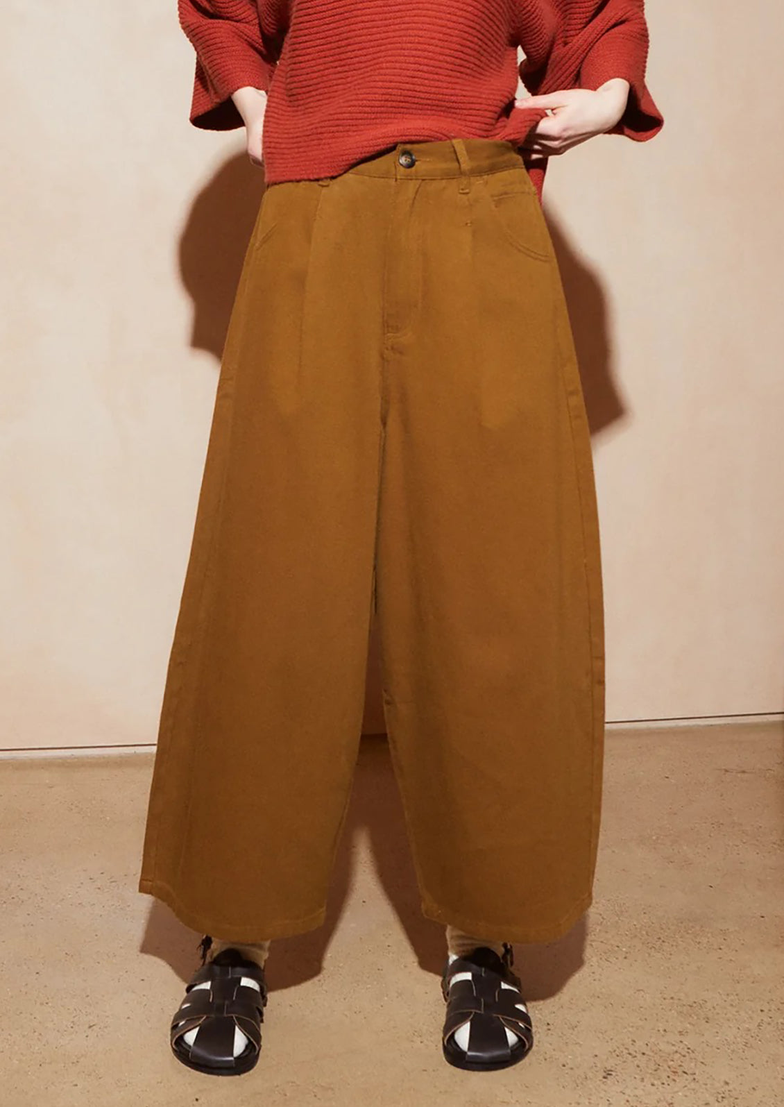 A person wearing wide-leg brown trousers, a rust-red ribbed sweater, and black sandals, standing against a plain beige wall.